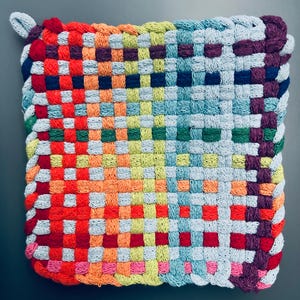 May include: A square, woven potholder in a rainbow of colors. The potholder features a loop for hanging and is made of a tightly woven fabric. The colors include red, orange, yellow, green, blue, and purple, creating a vibrant and cheerful design.