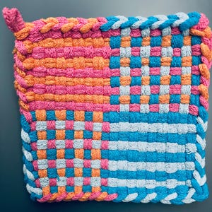May include: A square, woven hot pad in vibrant colors. The pad features a patchwork design with sections of pink, orange, blue, and white. The edges are finished with a braided trim in coordinating colors, and a loop for hanging.
