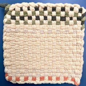May include: Woven square pot holder in cream, with a checkerboard pattern in green and black at the top. A band of peach-colored squares accents the bottom edge. The pot holder has a loop for hanging.