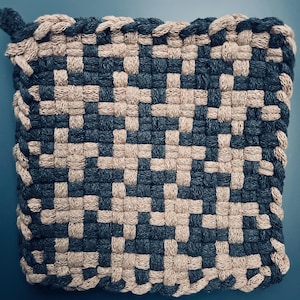 Könnte beinhalten: Quadratischer, gewebter Topflappen in einem Schachbrettmuster aus Marineblau und Beige. Die Kanten sind mit einer geflochtenen Bordüre in passender beiger Farbe versehen. Eine Schlaufe zum Aufhängen ist angebracht.