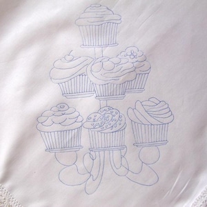 Puede incluir: Un mantel blanco con un diseño bordado en azul de un soporte para cupcakes con seis cupcakes. Los cupcakes tienen diferentes diseños de glaseado, incluyendo remolinos, puntos y una cereza en la parte superior.