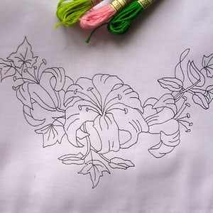 May include: White fabric with a black floral embroidery design of lilies and leaves. Three spools of embroidery thread in green, pink, and green are in the upper right corner. Suitable for embroidery projects.