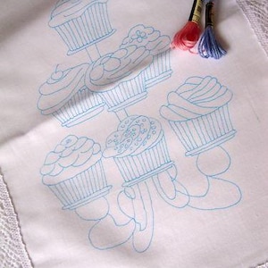 May include: A white embroidery design of a cupcake chandelier with six cupcakes, each with a different design. The cupcakes are outlined in blue thread and have a white background.