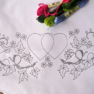 May include: A black and white line drawing of two intertwined hearts surrounded by a vine with leaves and flowers. The design is on a white fabric background.