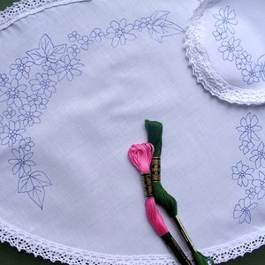 May include: White oval tablecloth with a crocheted border and blue embroidered floral designs. Two spools of embroidery floss, one pink and one green, are lying on the tablecloth.