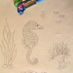Può includere: Un primo piano di un tessuto beige con un disegno a linea nera di un cavalluccio marino, alghe e coralli. Tre bobine di filo da ricamo si trovano nell'angolo in alto a destra dell'immagine. Il filo è verde, rosa e blu.