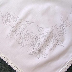 May include: White tablecloth with a delicate floral embroidery design. The embroidery features roses, leaves, and small flowers in a black thread. The tablecloth has a white lace trim around the edges.