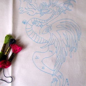 May include: A white fabric with a light blue dragon outline, ready for embroidery. Three spools of colorful embroidery floss in green, dark blue, and pink are visible, suggesting a craft project. The dragon design features intricate details.