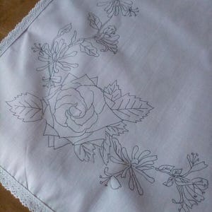 May include: White fabric with a black embroidered design of a rose and honeysuckle flowers. The fabric has a lace trim around the edges.