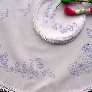 Könnte beinhalten: Weiße bestickte Tischdecke mit einem blauen Blumendesign, zwei Vögeln und einer Frau, die einen Blumenstrauß hält. Die Tischdecke hat einen Spitzenrand.