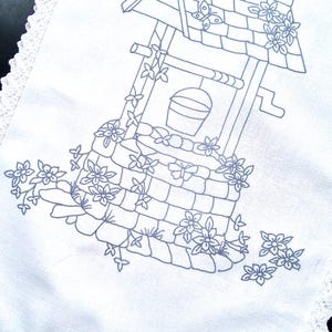 Peut inclure: Une nappe en lin blanc avec un motif brodé bleu d'un puits avec un seau, des fleurs et un papillon. La nappe a une bordure en dentelle.