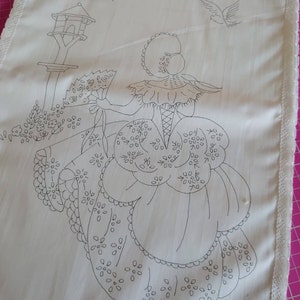 Puede incluir: Una tela blanca con un diseño de bordado negro de una mujer con un vestido sosteniendo un abanico, con pájaros volando sobre ella. El diseño incluye una casa para pájaros en un poste.