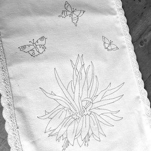 May include: A white linen tea towel with a lace border features an embroidered design of a flower with three butterflies. The flower is in the center of the towel and the butterflies are on either side of the flower.