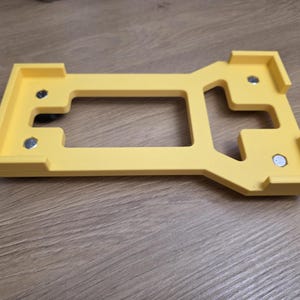 May include: A yellow, rectangular object with a central cutout and raised edges. The object has a unique shape with several right angles and appears to have small, silver fasteners. The item is likely a component or part of a larger assembly.