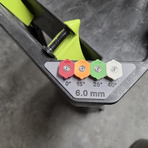 May include: A close-up of a pressure washer nozzle holder with four colored nozzles: red (0°), orange (15°), green (25°), and white (40°). The holder is gray with a yellow accent and the text "6.0 mm".