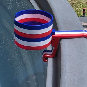 May include: A red, white, and blue striped cup holder, designed to clamp onto the edge of a pool or similar structure. The holder is made of plastic and features a patriotic color scheme.