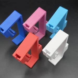May include: A collection of colorful phone stands in blue, pink, light blue, red, and white. Each stand has a rectangular base and a vertical support with a slot for holding a phone. The stands are made of a solid material.