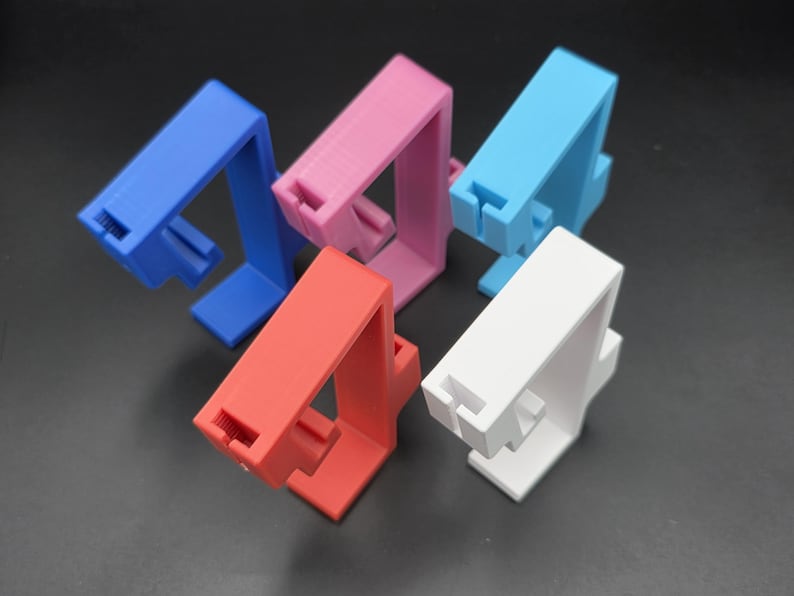 May include: A collection of colorful phone stands in blue, pink, light blue, red, and white. Each stand has a rectangular base and a vertical support with a slot for holding a phone. The stands are made of a solid material.