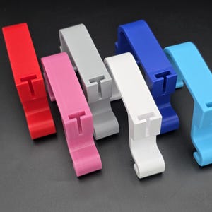 May include: A collection of colorful, plastic clips in red, pink, gray, white, blue, and light blue. Each clip has a T-shaped slot and a curved base. The clips are arranged on a dark surface.