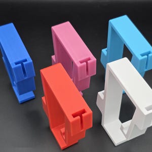 May include: Five colorful 3D-printed phone stands in blue, pink, red, light blue, and white. Each stand has a rectangular top with a T-shaped slot and a rectangular base. The stands are arranged on a dark surface.