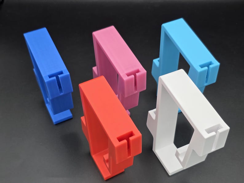 May include: Five colorful 3D-printed phone stands in blue, pink, red, light blue, and white. Each stand has a rectangular top with a T-shaped slot and a rectangular base. The stands are arranged on a dark surface.