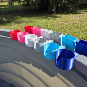 May include: A collection of colorful cup holders in various shades of red, pink, white, gray, light blue, and dark blue. The cup holders are designed to attach to the side of a surface.