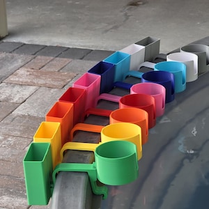 May include: A row of colorful cup holders in various shades, including green, yellow, orange, red, pink, purple, blue, and white. Each holder has a handle and is designed to attach to a surface. The holders are made of plastic.