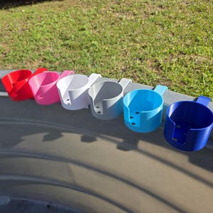 May include: A row of six colorful cup holders in red, pink, white, gray, light blue, and dark blue. Each holder has a handle and is designed to clip onto a surface. The holders are made of plastic.
