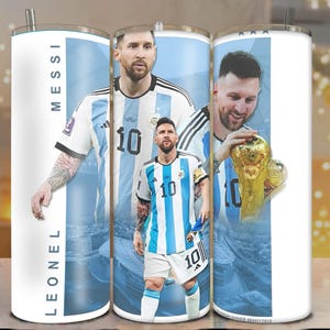 May include: A white stainless steel tumbler featuring images of Lionel Messi in his Argentina soccer uniform. The tumbler has a silver straw and the name "LEONEL MESSI" printed vertically. The design includes the number 10 and a gold trophy.