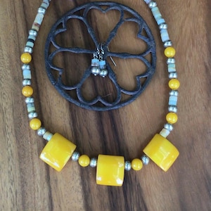 May include: A handmade jewelry set featuring a yellow and silver beaded necklace with square yellow beads. The set includes matching earrings and is displayed with a decorative metal piece and a purple flower on a wooden surface.