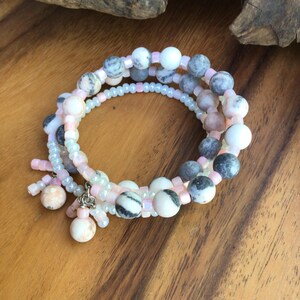 May include: A multi-strand wrap bracelet featuring a combination of round, variegated grey, white, and peach-toned stone beads interspersed with small, translucent pale pink seed beads. Two dangling bead charms hang from a silver-toned metal ring.
