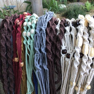 May include: A collection of macrame plant hangers made from various colored cotton cords including dark brown, maroon, mustard yellow, sage green, slate blue, and cream. Each hanger features intricate knotted patterns and wooden beads.