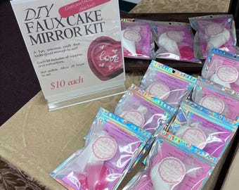Könnte beinhalten: Eine Präsentation von DIY Faux Cake Mirror Kits. Die Kits sind in transparenten, schillernden Reißverschlussbeuteln verpackt und enthalten Zubehör und Anweisungen. Ein Schild wirbt für das Handwerk, mit einem herzförmigen Kuchendesign und dem Wort "love".