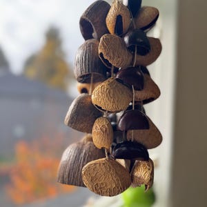 May include: A hanging wind chime crafted from various sizes of natural coconut shells. The shells are strung together with twine, showcasing a range of brown hues and textures. The chime hangs in front of a blurred outdoor scene.