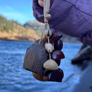 May include: A handcrafted wind chime featuring a variety of natural materials. The chime includes dark brown coconut shells, light brown seed pods, and dark red-brown acorn caps, all strung together with a beige cord. The background shows a body of water and mountains.
