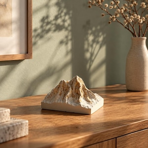 May include: A white and gold mountain sculpture with a textured, jagged peak design. The piece is mounted on a flat, rectangular base, showcasing detailed ridges and crevices that mimic a natural mountain range in a compact, decorative form.