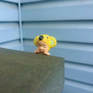 Puede incluir: Una peque&ntilde;a figura de un personaje con los ojos cerrados y las manos bajo la barbilla. El personaje lleva un gorro amarillo brillante y texturizado que recuerda a un koala, con una nariz oscura brillante y detalles iridiscentes.