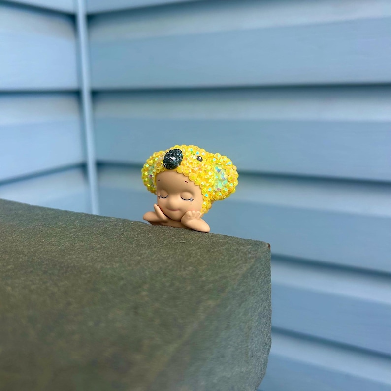 Puede incluir: Una peque&ntilde;a figura de un personaje con los ojos cerrados y las manos bajo la barbilla. El personaje lleva un gorro amarillo brillante y texturizado que recuerda a un koala, con una nariz oscura brillante y detalles iridiscentes.