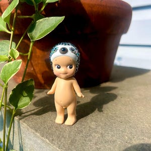 Puede incluir: Una peque&ntilde;a figura de piel clara con cabeza y cuerpo lisos y redondeados, que lleva un gorro blanco adornado con una capa densa de brillantes multicolores y un &uacute;nico acento circular oscuro centrado en la zona de la frente.