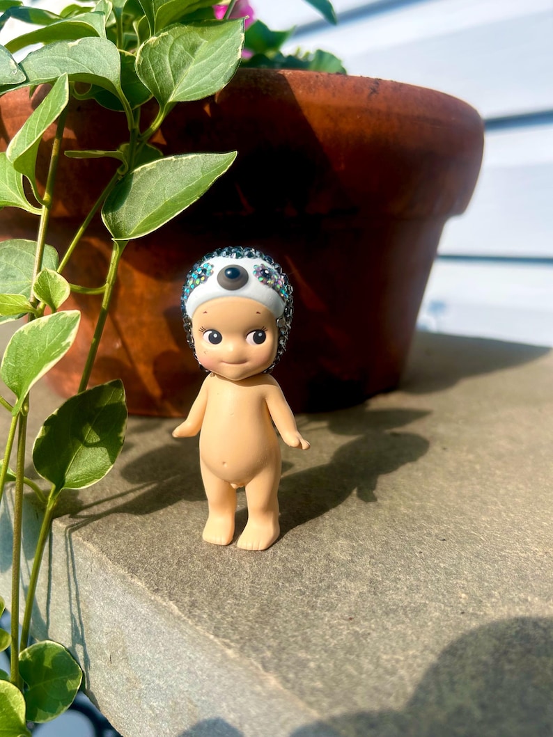 Puede incluir: Una peque&ntilde;a figura de piel clara con cabeza y cuerpo lisos y redondeados, que lleva un gorro blanco adornado con una capa densa de brillantes multicolores y un &uacute;nico acento circular oscuro centrado en la zona de la frente.