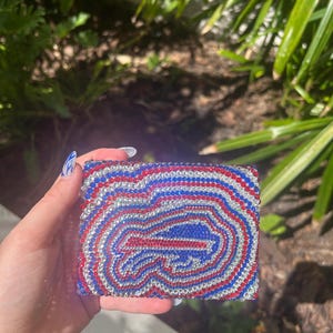 Puede incluir: Una cartera rectangular adornada con pedrería con un diseño rojo, blanco y azul. El diseño es un logotipo estilizado. La cartera está cubierta de pequeños y brillantes pedrería.