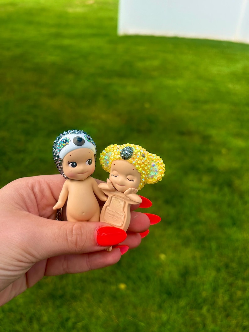 Puede incluir: Dos peque&ntilde;as figuras coleccionables con cuerpos lisos de color piel. Una figura lleva un tocado de perezoso incrustado con cristales azul gris&aacute;ceo, mientras que la otra presenta un tocado de koala amarillo brillante incrustado con cristales con las manos en la cara.