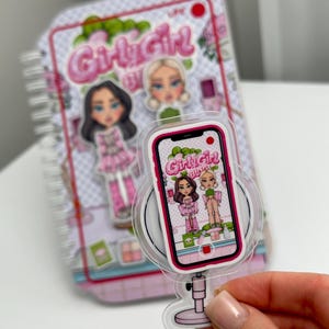Könnte beinhalten: Ein rosa-weißes Notizbuch zum Thema Girly Girl und eine transparente, acrylfarbene Dekoration in Form eines Telefons. Die Dekoration zeigt ein Ringlicht und einen Telefonbildschirm mit den Worten "Girly Girl Blogs" und Illustrationen von drei Mädchen.