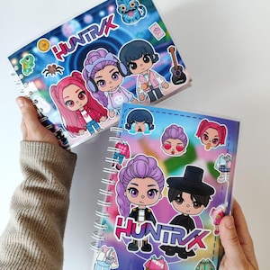 Può includere: Due quaderni a spirale con copertine colorate raffiguranti personaggi dei cartoni animati e la parola "HUNTRAX". Le copertine sono decorate con adesivi di vari disegni, tra cui un gufo, una chitarra e altre figure. I quaderni sono tenuti su uno sfondo bianco.
