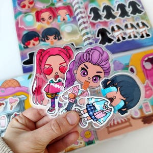 May include: A collection of colorful stickers featuring cartoon characters with various expressions. The stickers are in a clear plastic sheet, with a spiral-bound notebook in the background. The characters have pink, purple, and black hair.