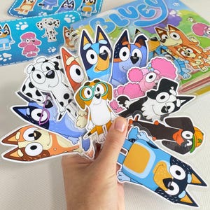 May include: A hand holding a selection of cartoon dog character stickers, including Bluey and Bingo. The stickers are brightly coloured and feature various breeds. A book and additional stickers are visible in the background.