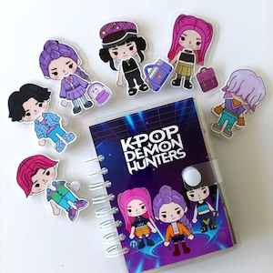 Peut inclure: Un carnet à spirales avec une couverture affichant "K-POP DEMON HUNTERS" et des illustrations de personnages de dessins animés. Plusieurs breloques en acrylique transparent des mêmes personnages sont disposées autour, chacune avec des tenues et coiffures différentes.