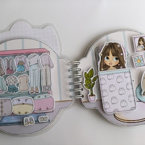 May include: A spiral-bound paper doll book with a wardrobe and bedroom scene. The wardrobe features paper doll clothing in various styles and colors. The bedroom scene includes a doll, a cat, and decorative items.