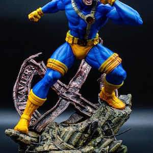 Peut inclure: Une figurine de collection détaillée représentant un super-héros en costume bleu et jaune avec une visière rouge, dans une pose dynamique sur un socle rocheux rempli de débris, incluant une structure métallique avec le mot XAVIER gravé.