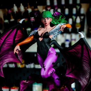 May include: A detailed figurine of a female character with green hair, black bodysuit, and purple leggings. The figure has large, dark purple wings and is posed dynamically above a swirling purple base. The figure's arms are orange and the wings have a pink interior.
